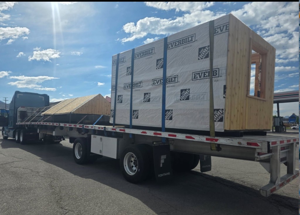 Semi truck with flatbed trailer carrying oversized modular wooden building sections secured with straps on a sunny day.