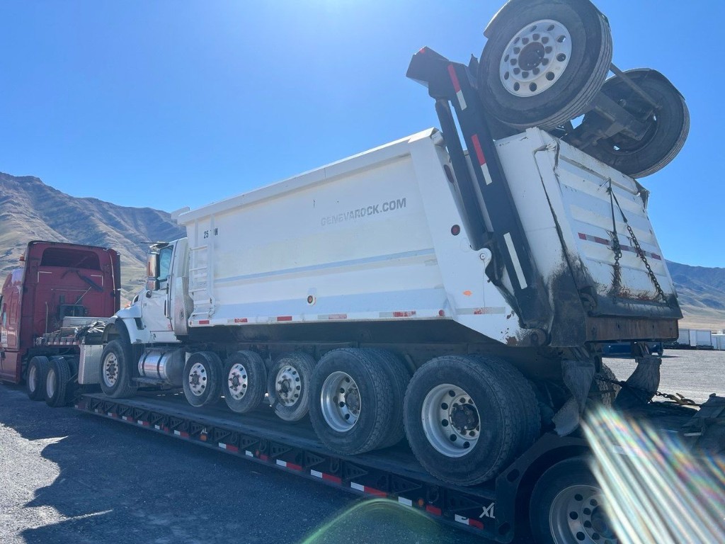 Heavy-duty dump truck secured on a multi-axle lowboy trailer with red tractor, open landscape and mountains in the background.