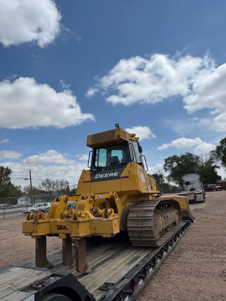 Yellow crawler dozer secured on a lowboy trailer, rear view with ripper and tracks visible under a partly cloudy sky.