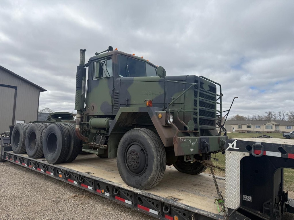Camouflage military-style heavy truck secured on a multi-axle lowboy trailer on a gravel lot.