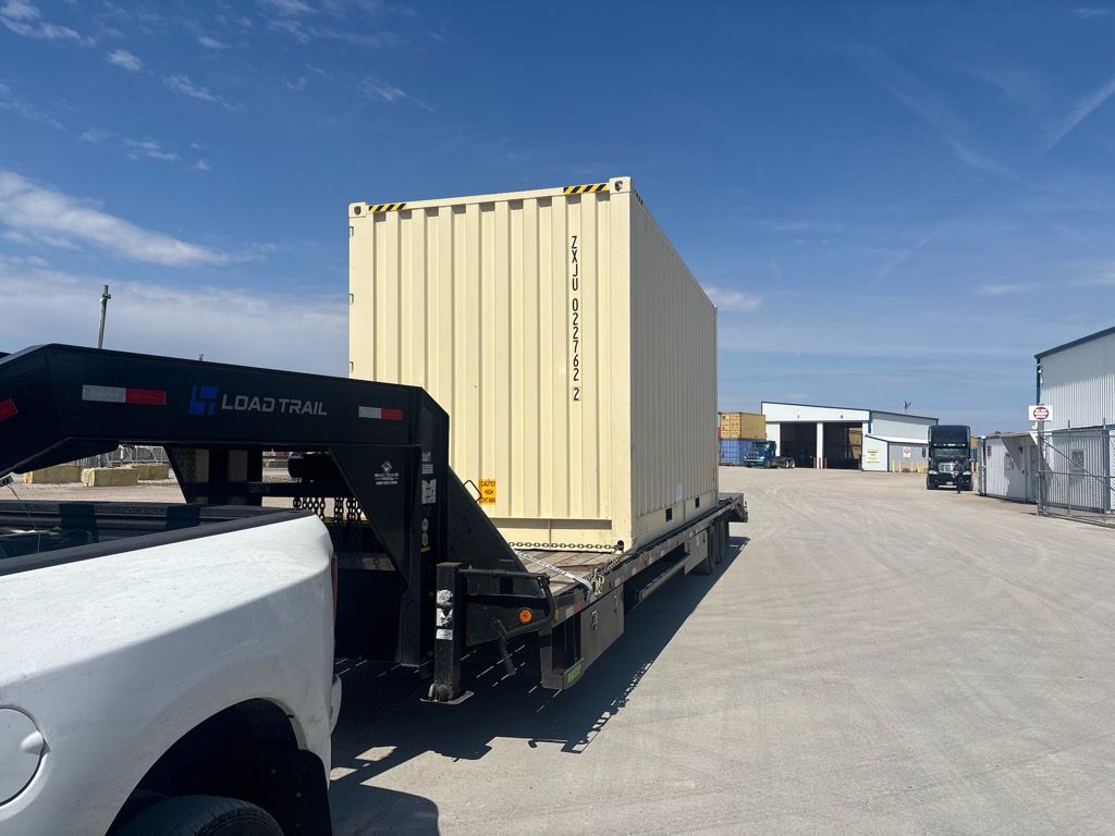 Shipping container secured on a black gooseneck flatbed trailer hitched to a white pickup truck in an industrial lot.